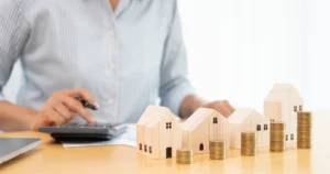 Person using a calculator beside a row of small model houses and coin stacks