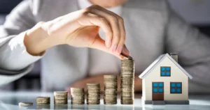Hand stacking coins next to a small model house