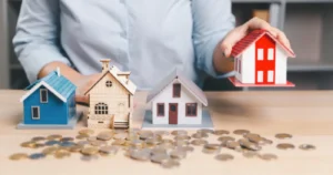 Four small model houses on a desk with scattered coins, one held under a hand
