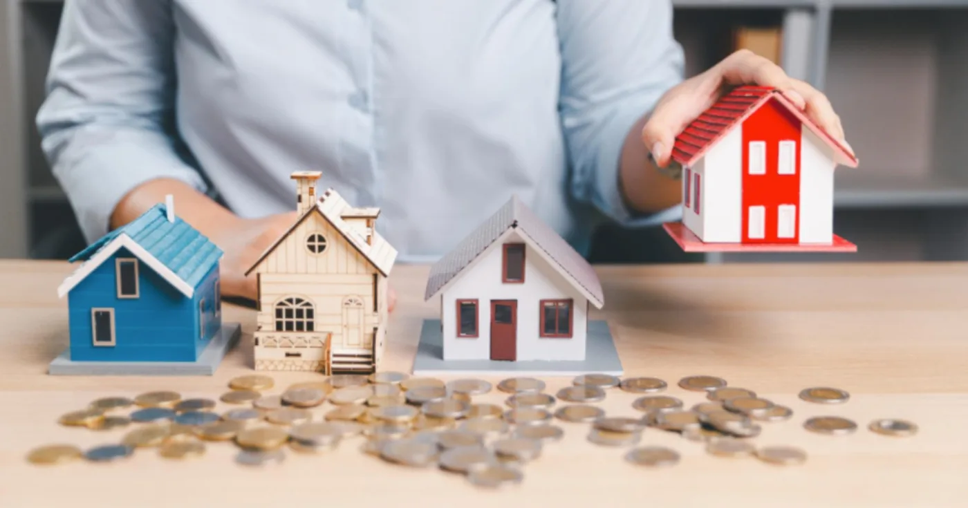Four small model houses on a desk with scattered coins, one held under a hand