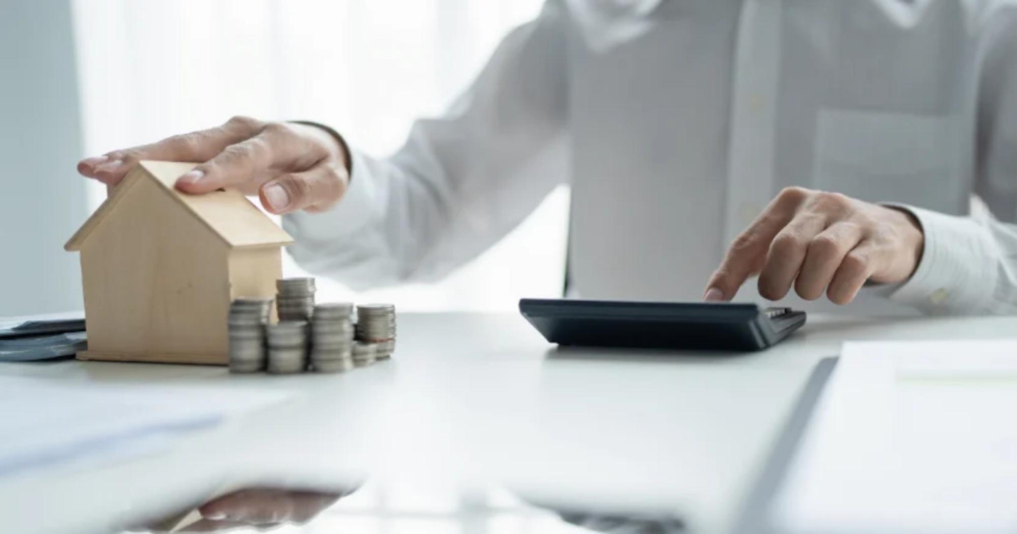 Hand resting on a small model house beside coin stacks while using a calculator