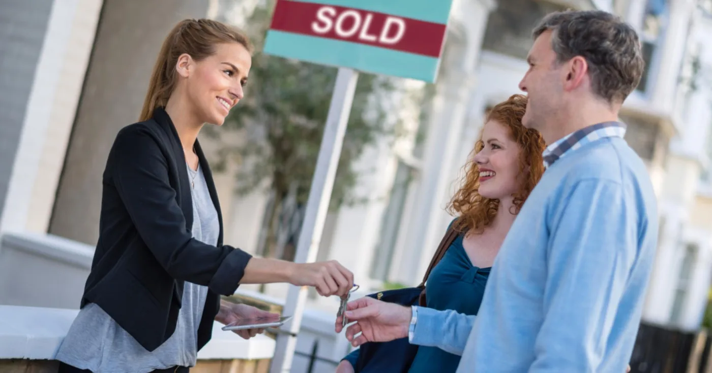 Agent handing keys to a smiling couple in front of a sold sign