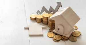 Wooden model house beside stacked gold coins with a rising arrow