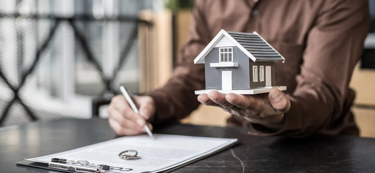 Person signing a document while holding a small model house